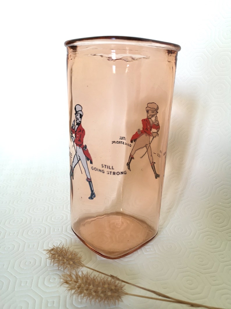 Peut inclure: Un verre en verre rose avec un design vintage repr&eacute;sentant deux hommes en veste rouge et pantalon blanc. Le texte "Still Going Strong" est imprim&eacute; sur le verre.