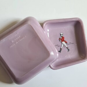 K&ouml;nnte beinhalten: Zwei quadratische, lavendelfarbene Aschenbecher aus Keramik. Ein Aschenbecher hat den Text "BLANGY MADE IN FRANCE" auf der Unterseite. Der andere Aschenbecher zeigt ein Schwarzwei&szlig;bild eines Mannes in roter Jacke und wei&szlig;er Hose, der geht. Der Text "JOHN WALKERS SONS SCOTCH WHISKY" ist unter dem Bild gedruckt.