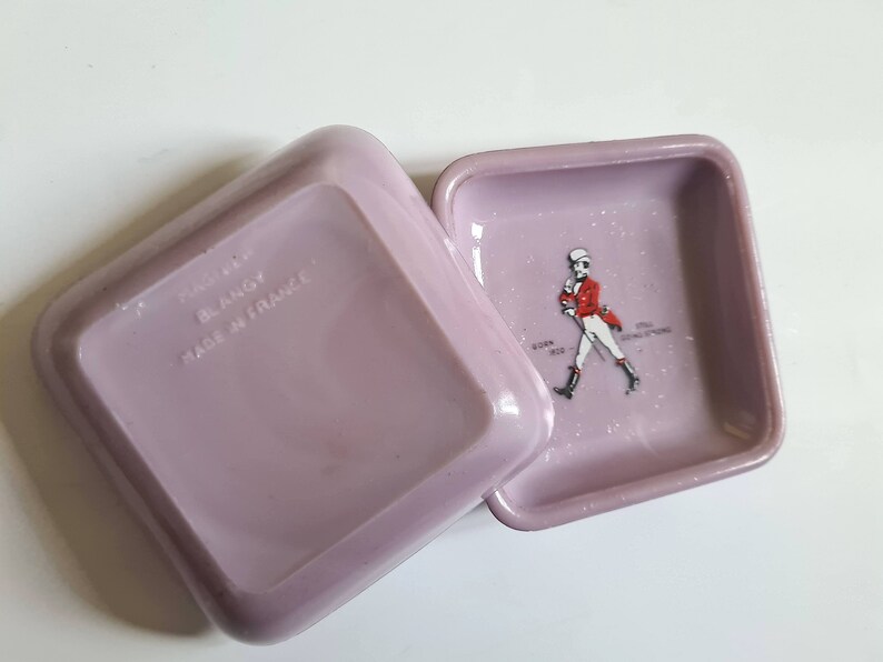 K&ouml;nnte beinhalten: Zwei quadratische, lavendelfarbene Aschenbecher aus Keramik. Ein Aschenbecher hat den Text "BLANGY MADE IN FRANCE" auf der Unterseite. Der andere Aschenbecher zeigt ein Schwarzwei&szlig;bild eines Mannes in roter Jacke und wei&szlig;er Hose, der geht. Der Text "JOHN WALKERS SONS SCOTCH WHISKY" ist unter dem Bild gedruckt.