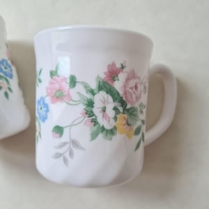 May include: White ceramic mug with a floral design featuring pink, blue, and yellow flowers with green leaves. The mug has a curved handle and a twisted base. The floral pattern is repeated around the mug.