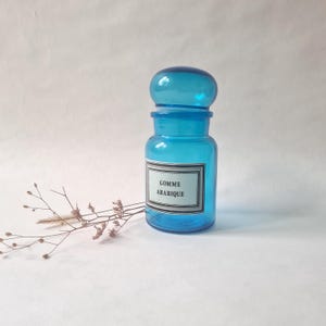 Peut inclure: Un pot de pharmacie en verre turquoise avec un couvercle arrondi. Le pot porte une étiquette avec le texte "GOMME ARABIQUE" dans un cadre noir. Une brindille de fleurs séchées repose près du pot.