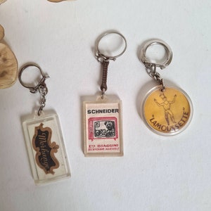 May include: Three vintage keychains with different designs. The first keychain features a clear plastic rectangle with a brown and white label that says "Moskovitch". The second keychain features a clear plastic rectangle with a black and white image of a television and the text "Schneider Ets Biaggini 81, Bd. Chave Marseille". The third keychain features a round, clear plastic keychain with a yellow background and a black and white image of a person in a yellow shirt and black shorts with the text "Lamoussette".
