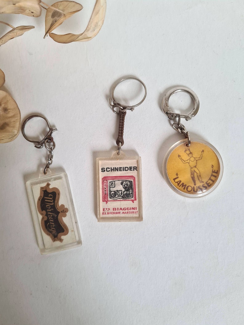 May include: Three vintage keychains with different designs. The first keychain features a clear plastic rectangle with a brown and white label that says "Moskovitch". The second keychain features a clear plastic rectangle with a black and white image of a television and the text "Schneider Ets Biaggini 81, Bd. Chave Marseille". The third keychain features a round, clear plastic keychain with a yellow background and a black and white image of a person in a yellow shirt and black shorts with the text "Lamoussette".