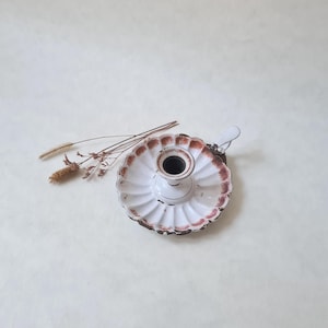 Puede incluir: Un candelero blanco de estilo vintage con un borde festoneado y un borde marrón óxido desgastado. El candelero tiene un asa pequeña y un pozo central para la vela. Se colocan hierbas secas cerca del candelero.