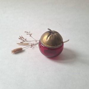 Peut inclure: Une boîte à bijoux décorative en forme de pomme. La base est en verre rouge foncé, surmontée d'un couvercle en métal doré. Une petite cuillère argentée repose sur le bord. Une brindille de fleurs séchées est à côté de la boîte.