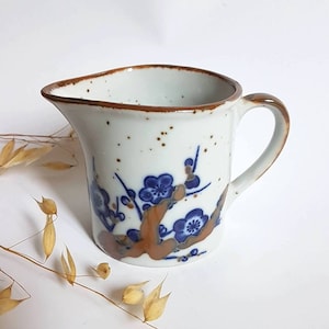 Op de afbeelding: Een klein wit keramiek kannetje met een bruine rand en een blauw bloemenmotief. Het kannetje heeft een handvat en een tuit.