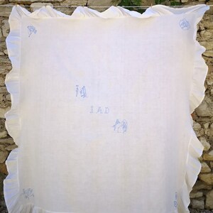 Puede incluir: Mantel de mesa de lino blanco con un dise&ntilde;o bordado en azul de una casa y personas. El mantel tiene un borde con volantes y la palabra "IMD" est&aacute; bordada en el centro.