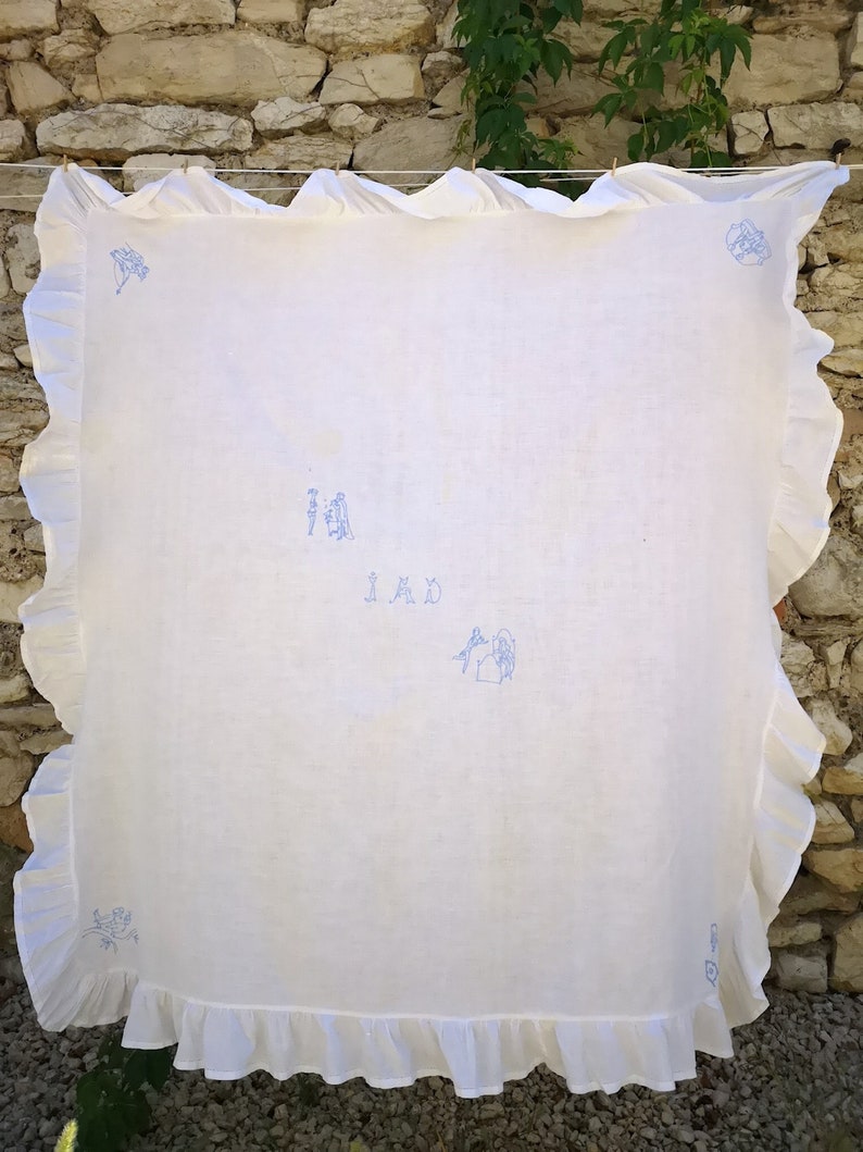 Puede incluir: Mantel de mesa de lino blanco con un dise&ntilde;o bordado en azul de una casa y personas. El mantel tiene un borde con volantes y la palabra "IMD" est&aacute; bordada en el centro.