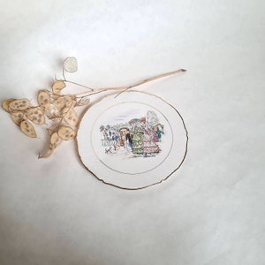 May include: A white ceramic plate with a gold rim features a colorful illustration of people gathered at an outdoor event. The scene includes umbrellas, hats, and a dog.