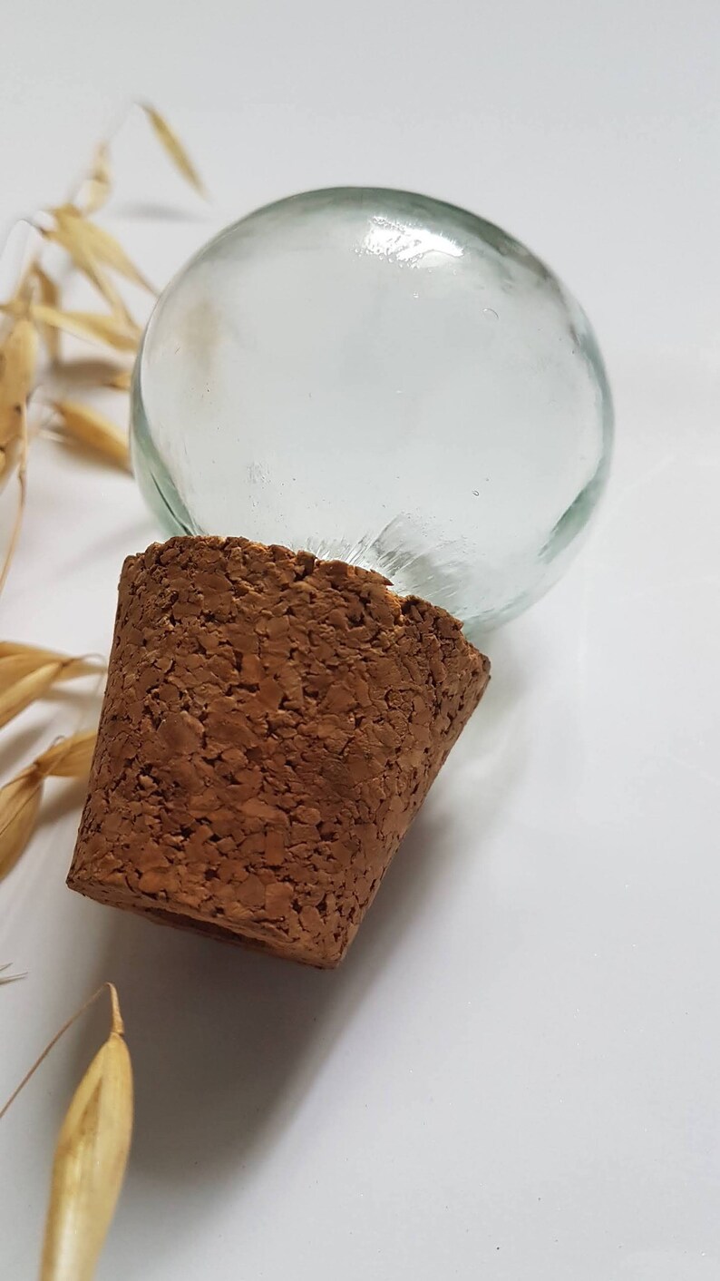 May include: A clear glass bottle stopper with a cork base. The bottle stopper is round and has a smooth surface.