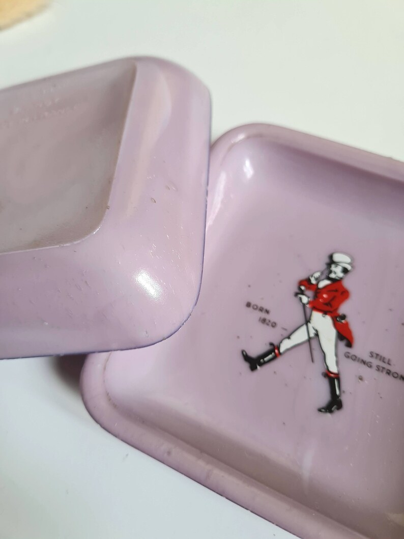 K&ouml;nnte beinhalten: Ein quadratischer Aschenbecher in Lila mit wei&szlig;em Rand. Der Aschenbecher zeigt ein Schwarzwei&szlig;bild eines Mannes in roter Jacke und wei&szlig;er Hose, der mit einem Stock geht. Der Text "Born 1920" und "Still Going Strong" ist auf dem Aschenbecher aufgedruckt.