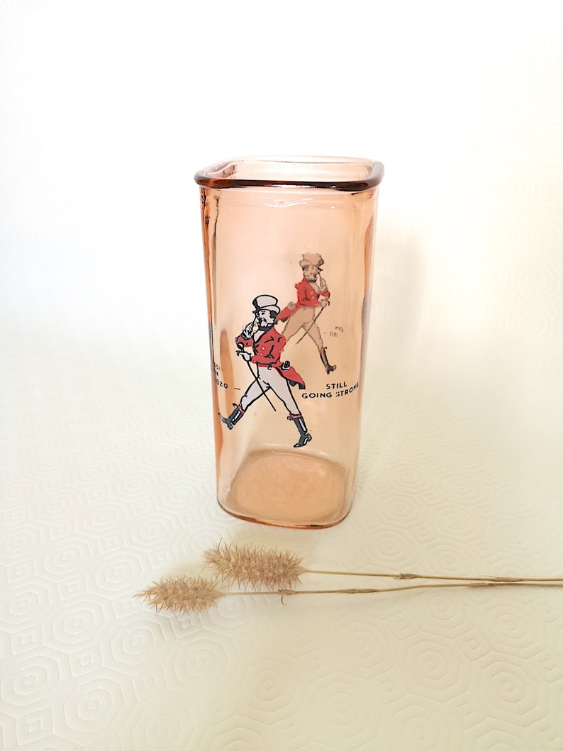 Peut inclure: Un verre &agrave; boire en verre rose avec une forme carr&eacute;e et une image imprim&eacute;e de deux hommes en v&ecirc;tements rouge et noir. Le texte "Still Going Strong" est imprim&eacute; sur le verre.