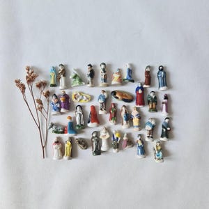 May include: A collection of thirty-five miniature ceramic figurines depicting various figures, including people in colorful clothing, animals, and a baby in a manger, arranged alongside a single dried brown plant stem.