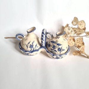 May include: A white ceramic oil and vinegar set with blue floral designs. The set has two small jugs with handles and a central holder with a handle.