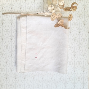 May include: A white linen tea towel with a red embroidered initial "M" on a white background. The towel is folded and placed on a patterned surface.