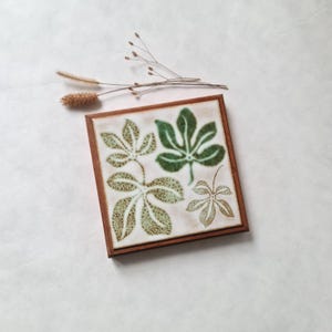 Peut inclure: Carreau de céramique carré avec un cadre en bois. Le carreau présente un motif de feuilles dans des tons de vert et de marron. Le carreau est carré, chaque côté mesurant environ 15 cm. Des brins séchés sont en arrière-plan.