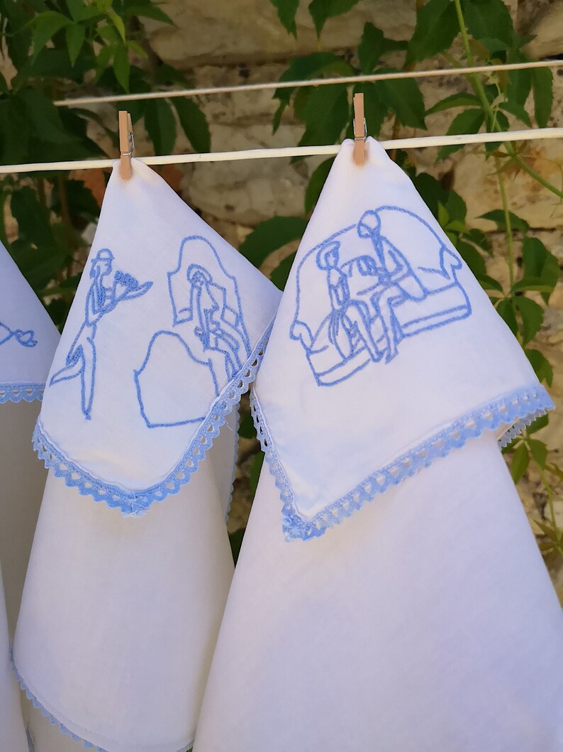 Puede incluir: Tres toallas de t&eacute; de lino blanco con dise&ntilde;os bordados en azul. Los dise&ntilde;os presentan personas en varias escenas. Las toallas tienen un borde de crochet azul.