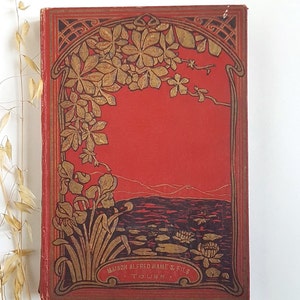 Op de afbeelding: Een rode boekomslag met een gouden en zwarte decoratieve rand. De rand heeft een bloemmotief met bladeren en bloemen. De titel van het boek is "Maison Alfred Mame & Fils Tours".