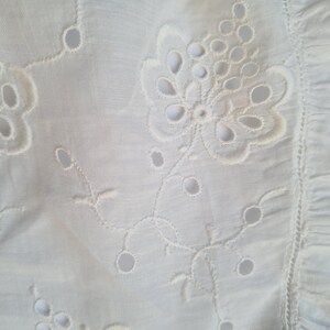May include: White fabric with a delicate embroidered floral pattern. The design features a large flower with smaller flowers and leaves surrounding it.
