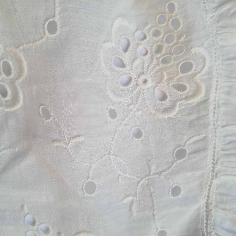 May include: White fabric with a delicate embroidered floral pattern. The design features a large flower with smaller flowers and leaves surrounding it.