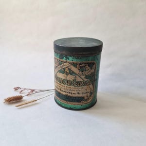 Op de afbeelding: Vintage cilindrische blikken doos met een donkergroen deksel en een turquoise body. De blikken doos heeft een label met sierlijke letters en bloemmotieven. De tekst op het label luidt "Pastilles Cristal" en andere Franse tekst. De blikken doos heeft een verouderd uiterlijk.