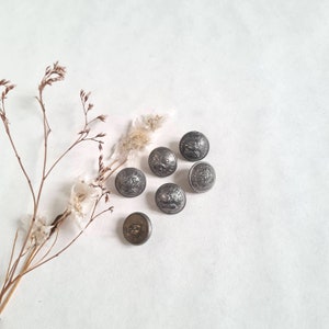 Peut inclure: Six boutons argentés avec un motif en relief. Les boutons sont disposés en demi-cercle sur un fond blanc.