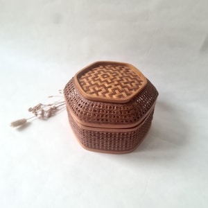Peut inclure: Une boîte hexagonale avec couvercle, fabriquée en rotin tressé marron avec un motif géométrique sur le dessus. Le récipient présente une base structurée et un couvercle amovible assorti, mettant en valeur des techniques de tressage naturel.