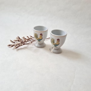 Puede incluir: Dos hueveras de cerámica blanca con diseño de gallo. Cada taza tiene un borde y una base dorados, y una ilustración colorida de gallo. Las hueveras están sobre una superficie blanca, con una ramita de flores secas a la izquierda.