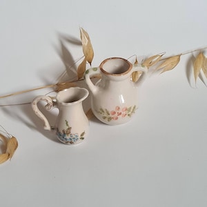 Op de afbeelding: Twee kleine, gebroken witte keramische kruiken met bloemmotieven. Eén kruik heeft een handvat en blauwe bloemen, de andere heeft twee handvatten en roze bloemen. Beide hebben een rustieke, handgemaakte uitstraling.