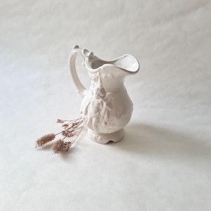 Peut inclure: Pichet en céramique blanche avec un motif orné de fleurs en relief. Le pichet a une anse et un bec.