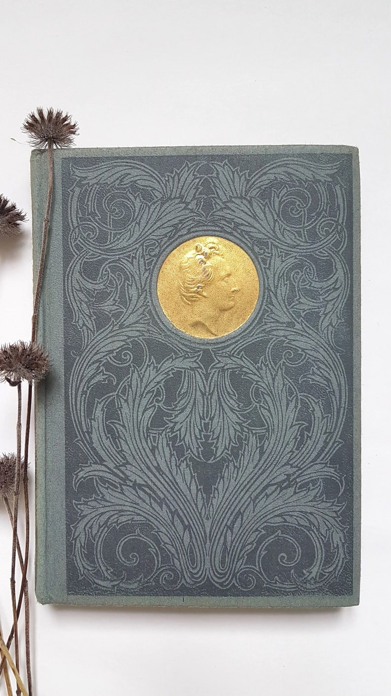 May include: A vintage book with a green cover and a gold embossed design. The design features a circular medallion with a profile of a person inside.