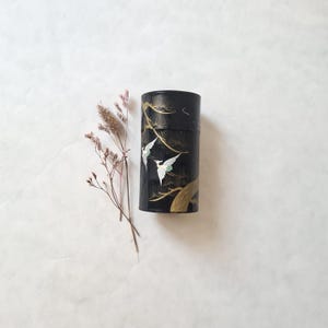 May include: Black cylindrical container with a gold and white design of birds and branches. The container is on a white surface with dried pink flowers.