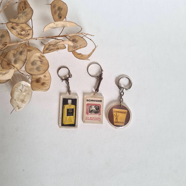 May include: Three keychains with different designs. The first keychain has a clear plastic charm with a bottle of cologne and the text "Eau de Cologne" on it. The second keychain has a plastic charm with the text "Schneider" and a picture of a television. The third keychain has a plastic charm with the text "Cristalor" and a picture of a man holding a bucket.