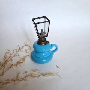 May include: A small blue glass oil lamp with a metal cage and a handle. The lamp is on a white surface.