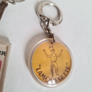 May include: A yellow round keychain with a clear plastic cover. The keychain features a cartoon illustration of a person wearing a hat and holding their arms out. The text "LAMOUSSETTE" is printed in brown around the edge of the keychain.