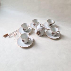 Peut inclure: Service à thé en porcelaine blanche avec six tasses et soucoupes, et un sucrier. Chaque pièce présente un motif et une bordure marron et dorés. L'ensemble est disposé sur une surface blanche.