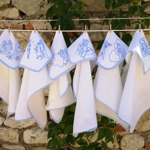 Puede incluir: Seis toallas de cocina blancas con bordados azules colgadas de un tendedero. Cada toalla presenta un dise&ntilde;o diferente de personas y animales.