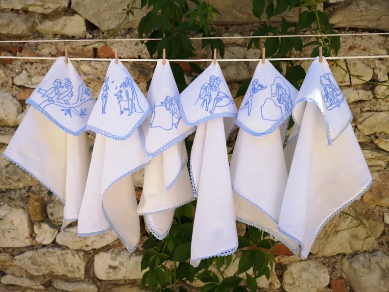 Puede incluir: Seis toallas de cocina blancas con bordados azules colgadas de un tendedero. Cada toalla presenta un dise&ntilde;o diferente de personas y animales.