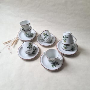 May include: Set of six white porcelain teacups and saucers with a floral design. Each cup and saucer features a different floral motif with green leaves and yellow flowers.
