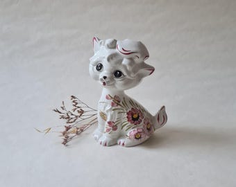 Puede incluir: Figura de cerámica blanca de un gato con detalles florales rosas y verdes. El gato está sentado con la cabeza inclinada hacia un lado y tiene una expresión caprichosa.