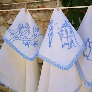 Puede incluir: Tres toallas de cocina blancas con bordados azules colgadas de una cuerda para tender la ropa. El bordado representa un p&aacute;jaro en una rama, dos figuras y un dise&ntilde;o floral.