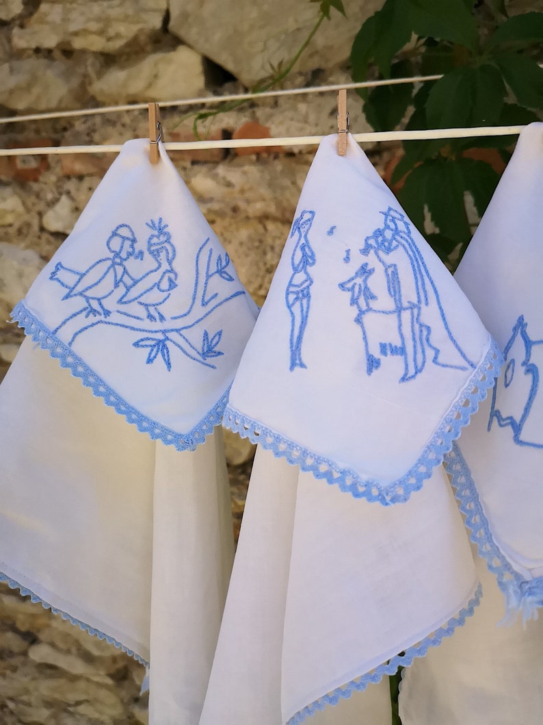 Puede incluir: Tres toallas de cocina blancas con bordados azules colgadas de una cuerda para tender la ropa. El bordado representa un p&aacute;jaro en una rama, dos figuras y un dise&ntilde;o floral.