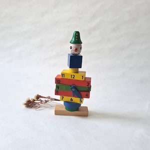 Puede incluir: Un payaso de madera de colores con un sombrero verde y una nariz roja. El payaso está formado por bloques apilados con los números del 1 al 12 impresos en ellos.