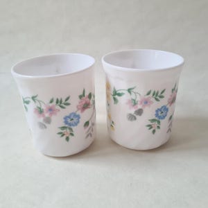 May include: Two white ceramic cups with floral designs. The cups feature a pattern of pink, blue, and yellow flowers with green leaves. The cups are cylindrical with a slightly flared top.