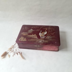 May include: A burgundy rectangular box with a detailed illustration of a crane in a landscape. The box has a keyhole and is likely made of wood. The artwork features gold and white accents, creating a vintage aesthetic.