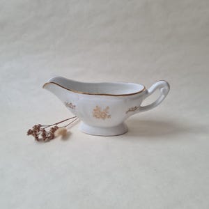 Peut inclure: Un saucier en céramique blanche avec un bord et une poignée dorés. Le saucier présente un motif floral doré et repose sur une petite base en piédestal. Quelques fleurs séchées sont au premier plan.