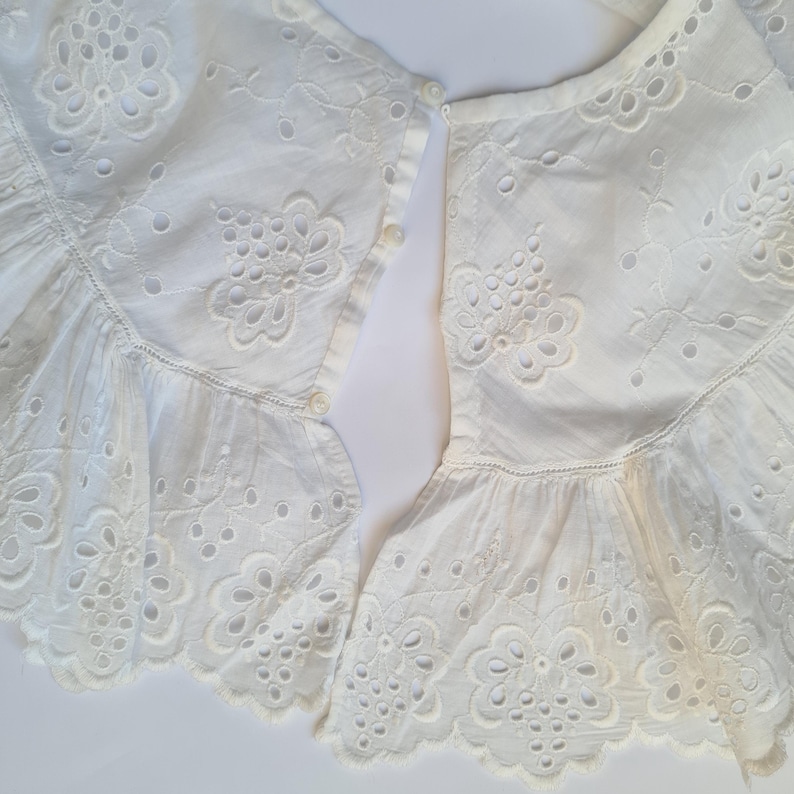 May include: Two white cotton lace collars with eyelet embroidery and a button closure. The collars are ruffled and have a delicate floral design.