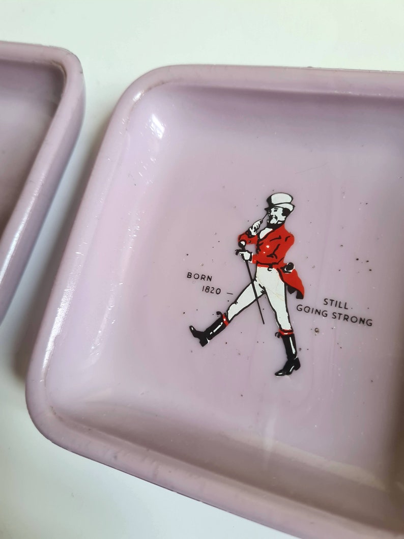 K&ouml;nnte beinhalten: Ein rosa Keramik-Aschenbecher mit einer Grafik eines Mannes in roter Jacke und Zylinder. Der Text "BORN 1820 STILL GOING STRONG" ist unter dem Mann gedruckt.