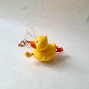 May include: A bright yellow rubber duck toy featuring a vibrant orange beak and orange webbed feet. The duck has white eyes with black pupils and a small yellow wing on its side. A small sprig of dried brown decorative grass is positioned behind it.