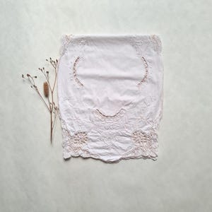 May include: A white, rectangular vintage tablecloth with detailed floral embroidery and a scalloped edge. The tablecloth has a central embroidered design and delicate lace details. A dried plant stem is placed on the side.